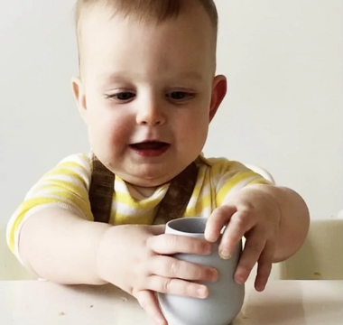 Baby holding gray silicone cup, mess-free mealtime with easy clean food catcher mat