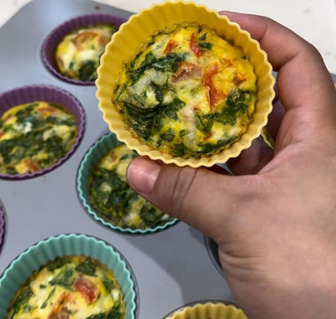 Hand holding yellow silicone egg muffin cup with spinach and tomato, baked in muffin tin
