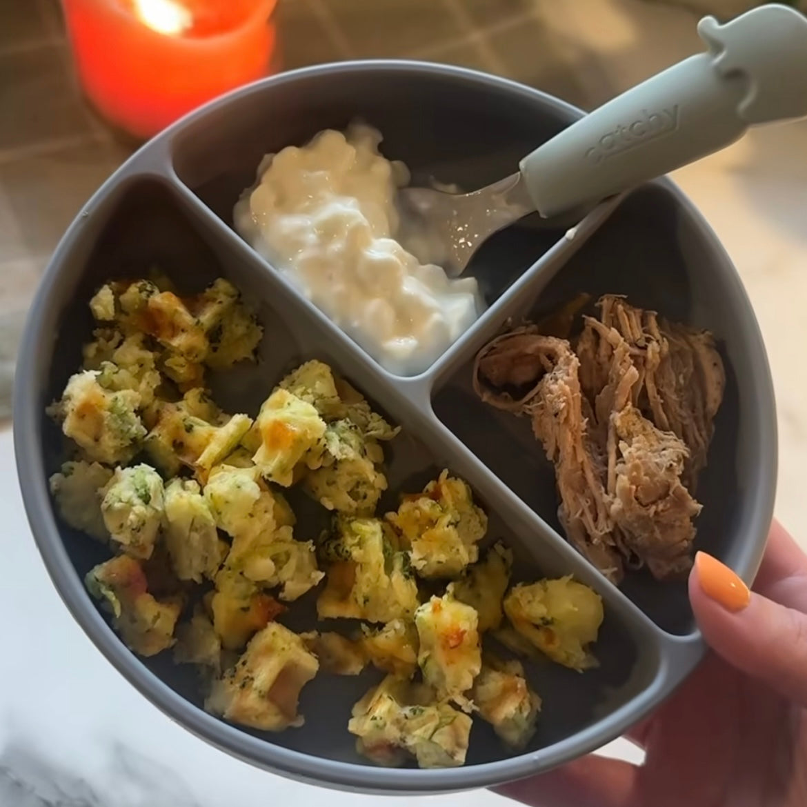 Gray divided baby plate with broccoli bites, yogurt, and shredded meat, held by hand with orange nail polish.