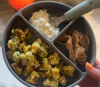 Gray divided baby plate with broccoli bites, yogurt, and shredded meat, held by hand with orange nail polish.