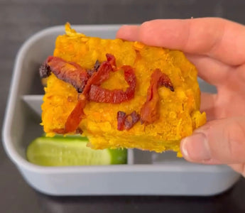 Hand holding savory yellow baked dish with sun-dried tomatoes over bento box with cucumber slices