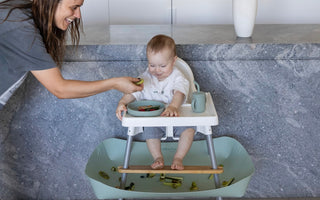 Baby in high chair with mess-free food catcher, mom feeding with smile